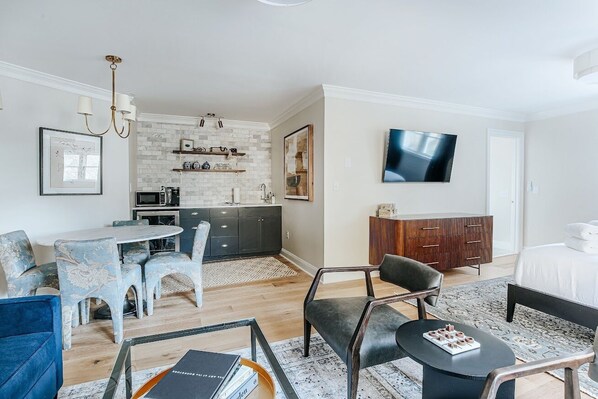 Una habitación de hotel moderna con zona de comedor, kitchenette y televisor montado en la pared.