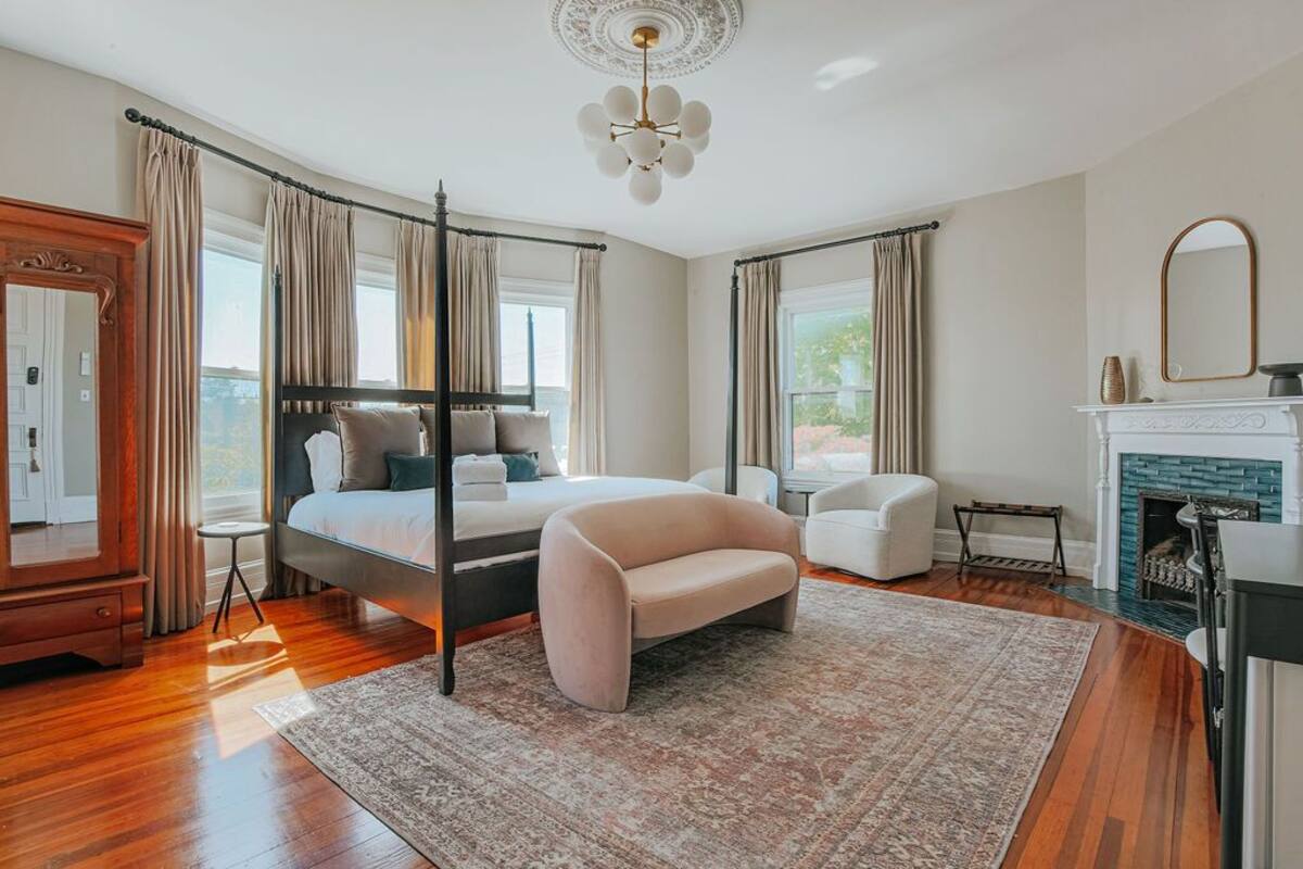 A bedroom with a four-poster bed, a sitting area with a sofa, a fireplace, and a large window.