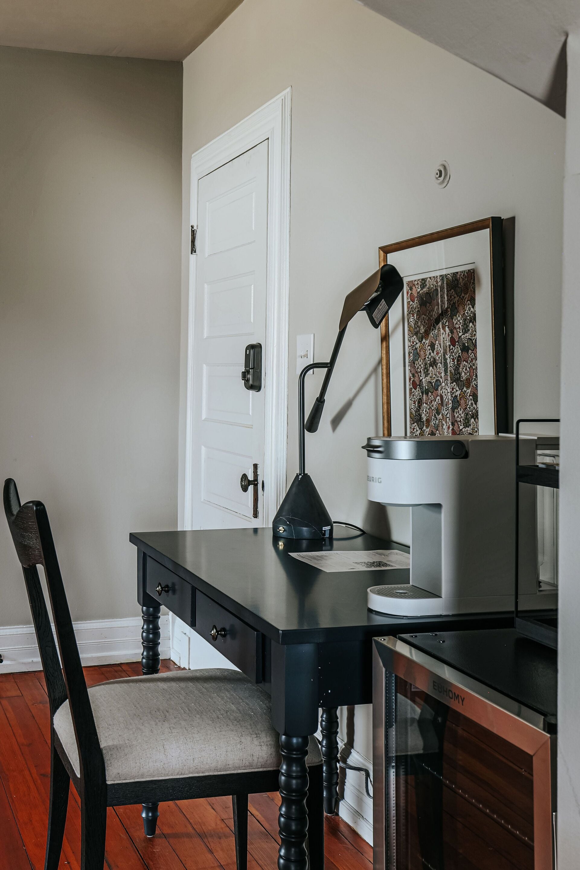 A room with a wooden chair, a desk with a lamp, a coffee maker, and a framed picture on the wall.