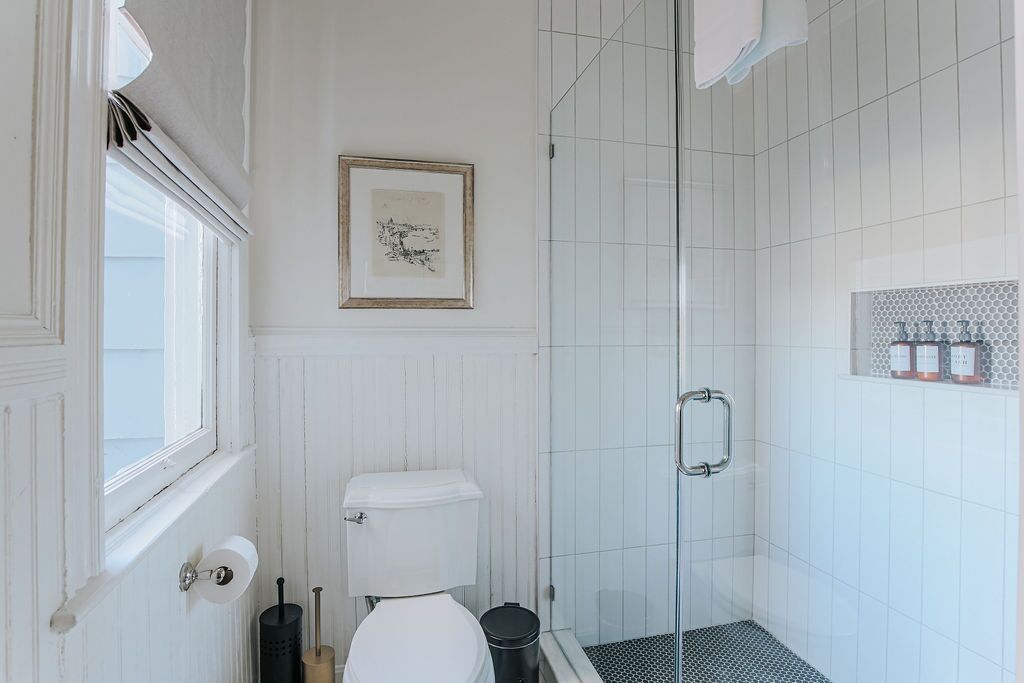 Ein Badezimmer mit einer weißen Toilette, einem gerahmten Kunstwerk an der Wand, einer Duschkabine mit Glastür und einem gefliesten Duschbereich.