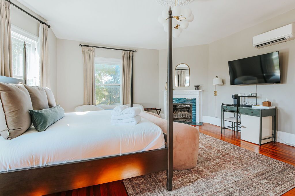 A bedroom with a large bed, a desk, a television, and a fireplace.