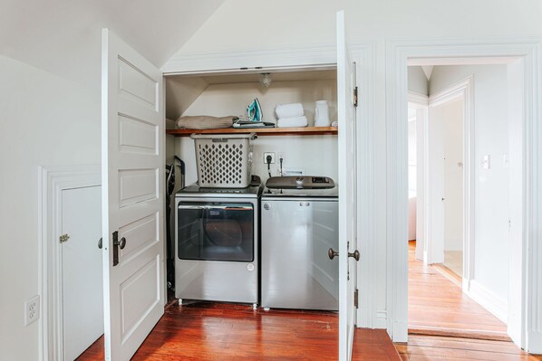 A compact kitchen with a built-in oven and dishwasher, a drying rack, and a shelf with towels and a kettle.