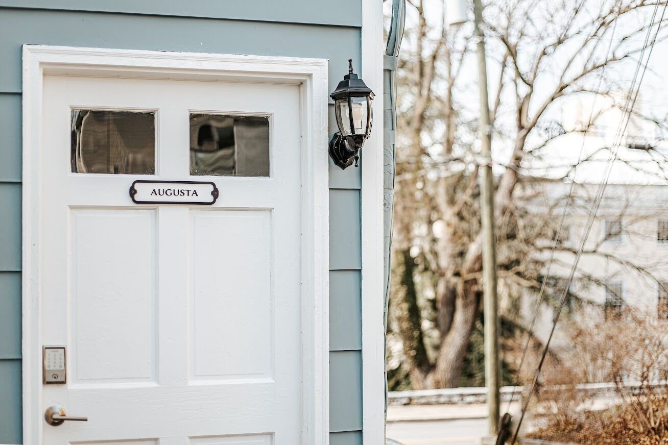 A white door with the name "AUGUSTA" above it and a black wall-mounted light fixture to the right.