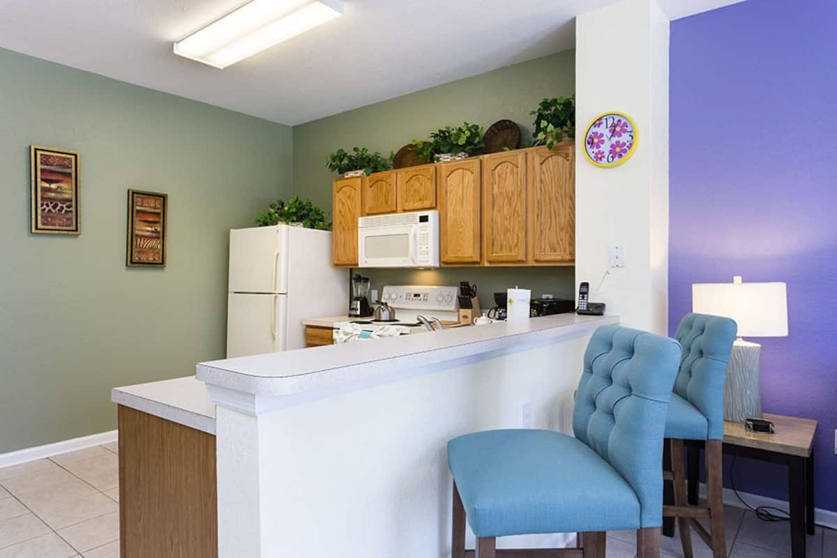 A kitchen with a central island, wooden cabinets, a white refrigerator, a microwave, and a sink. There are two blue chairs and a small table with a lamp.
