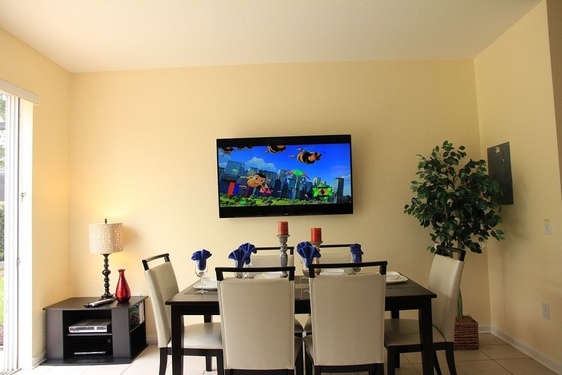 Un coin repas avec une table dressée pour quatre personnes, un téléviseur à écran plat fixé au mur, une petite table d’appoint avec une lampe et une plante en pot.