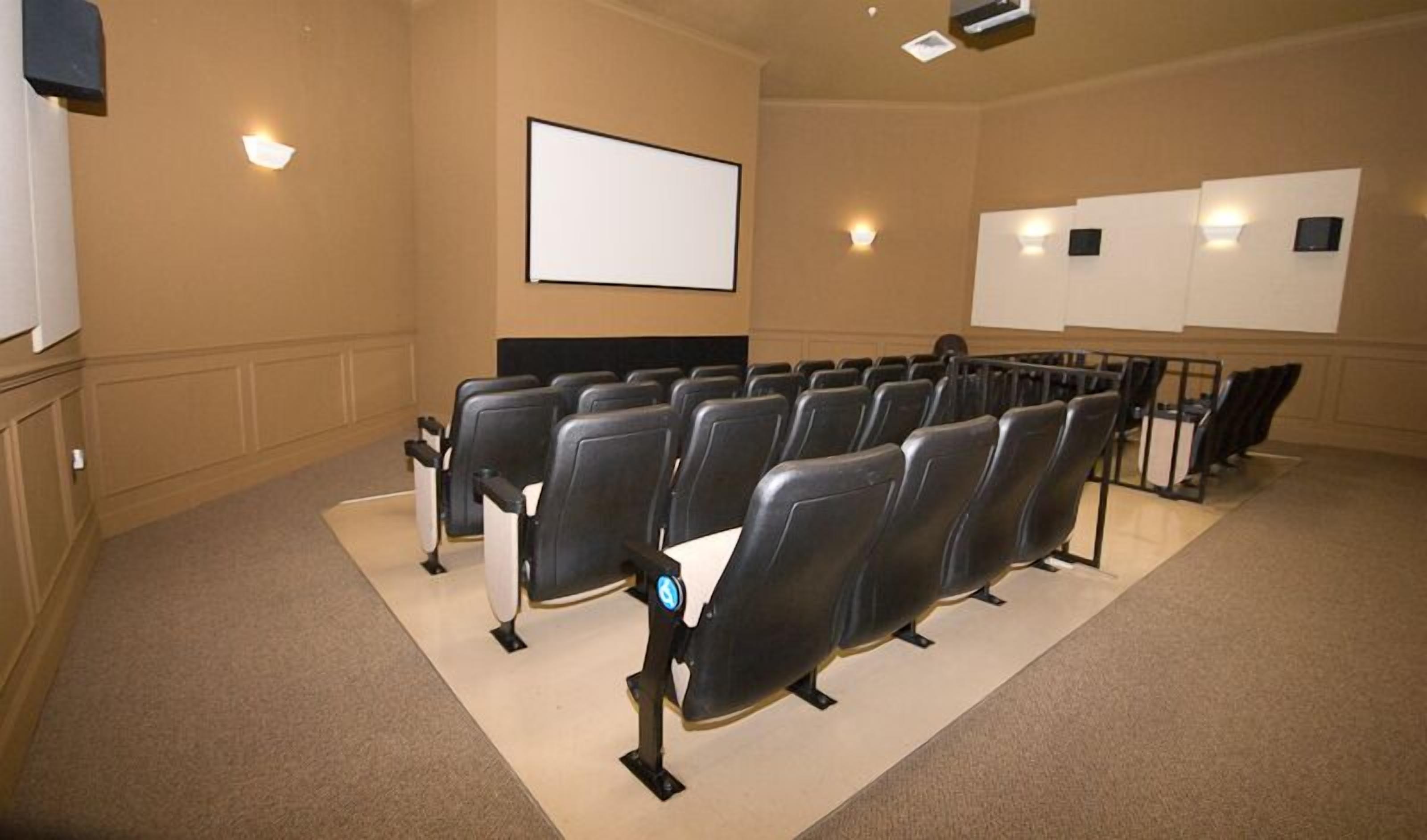 A conference room with rows of black chairs, a white projection screen, and wall-mounted speakers.