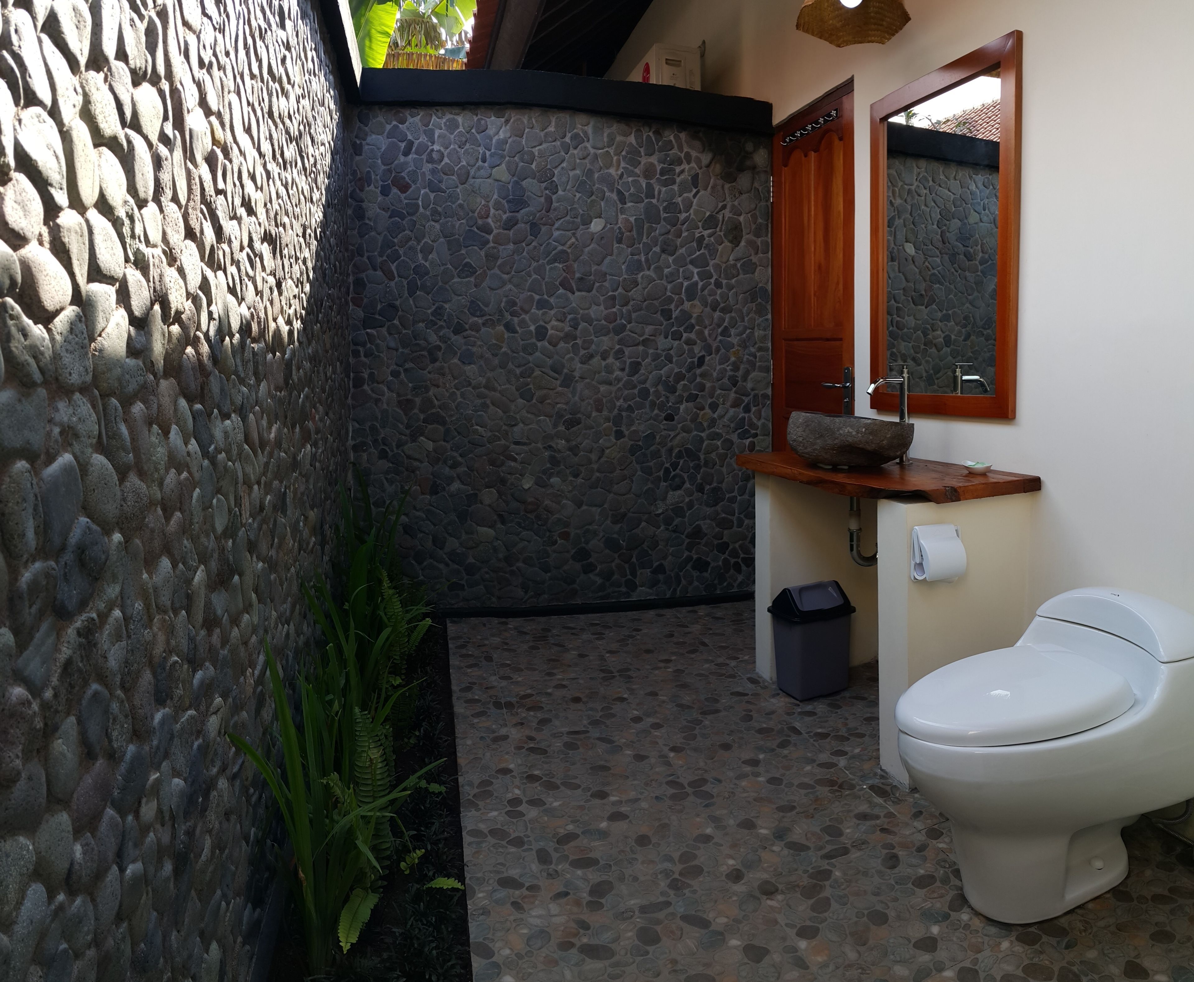 A bathroom with a stone wall, a wooden sink with a stone basin, a toilet, and a mirror above the sink.