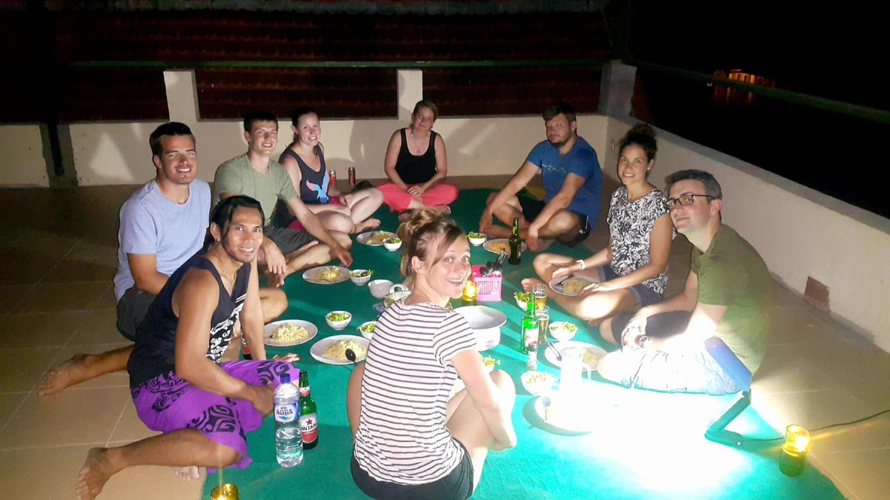Een groep mensen zit rond een tafel met eten en drinken, in een open setting met een betegelde vloer en een lage muur op de achtergrond.