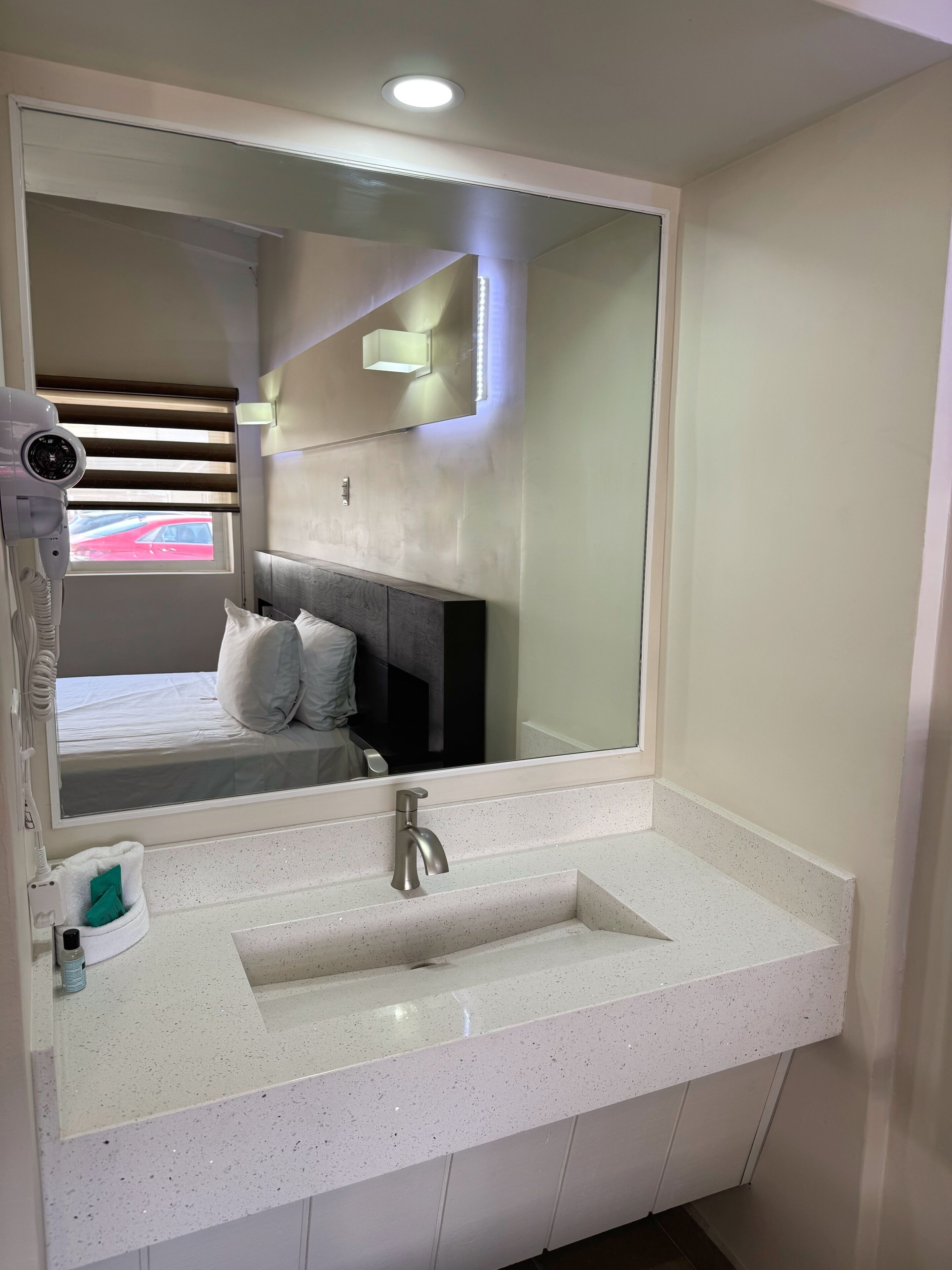 A hotel bathroom with a large mirror, a white sink, a hairdryer, and a bed with pillows.