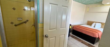 A hotel room with a shower, a bed with a red blanket, and a white door.