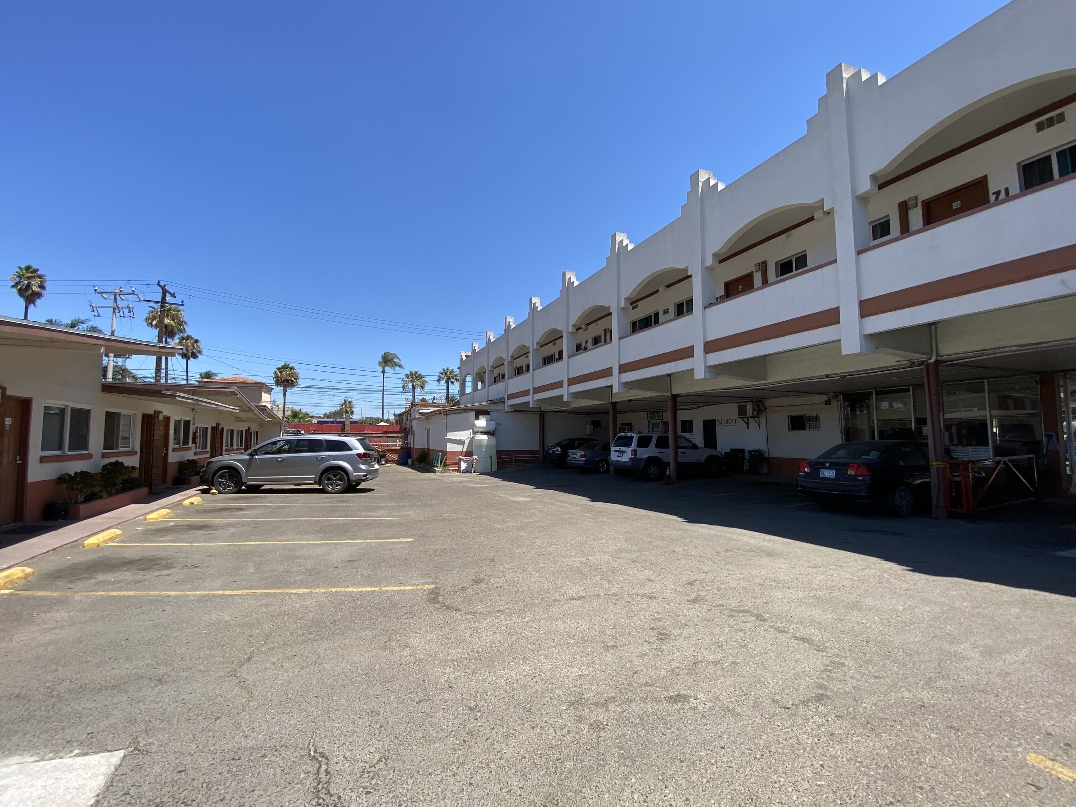 A parking lot with several cars, a building with multiple floors and balconies, and a clear blue sky.
