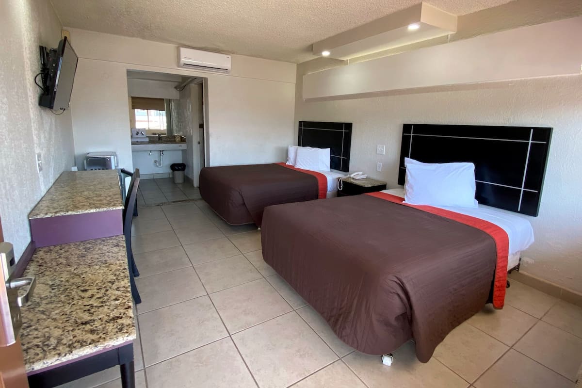 A hotel room with two beds, a small kitchenette, a TV mounted on the wall, and an air conditioning unit.