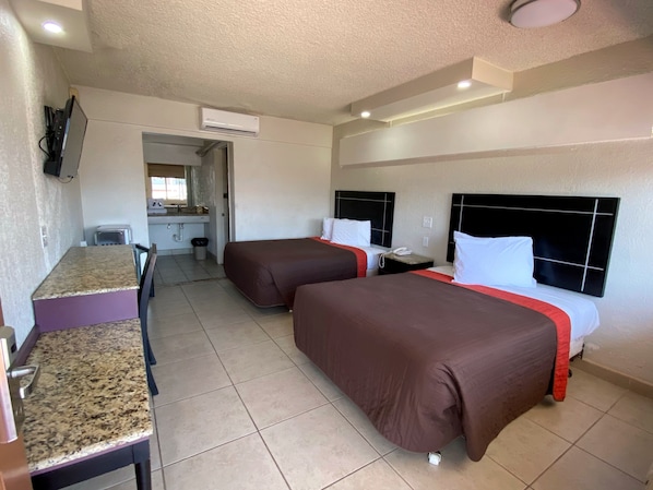 A hotel room with two beds, a small kitchenette, a TV mounted on the wall, and an air conditioning unit.