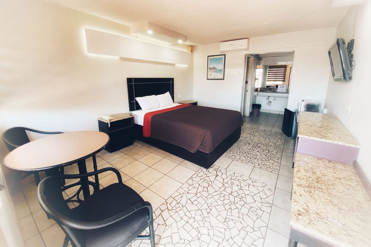 A hotel room with a bed, a desk, a chair, a small table, and a kitchenette.