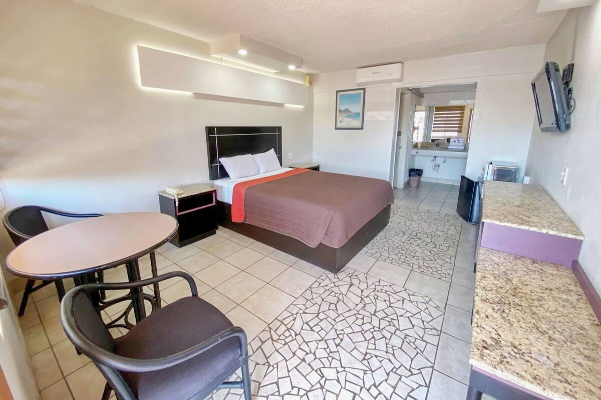 A hotel room with a bed, a desk, a chair, a small table, and a kitchenette.