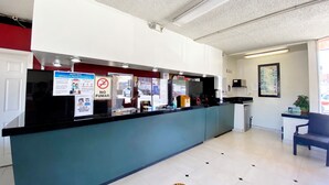 Een receptiebalie met een blauw aanrecht, witte muren en een klok. Er staan borden met preventiemaatregelen voor COVID-19 en een bord dat roken verbiedt.