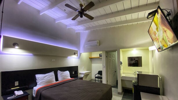 Een hotelkamer met een bed, een bureau, een stoel, een plafondventilator en een aan de muur bevestigde televisie.