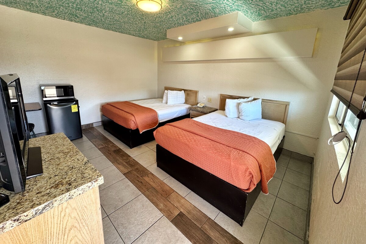 A hotel room with two beds, a kitchenette, a trash bin, and a ceiling light fixture.