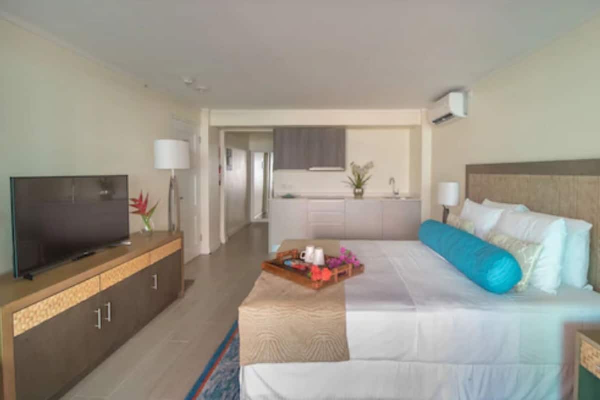 A hotel room with a bed, a TV on a wooden cabinet, a small kitchen area, and a bathroom visible through an open door.
