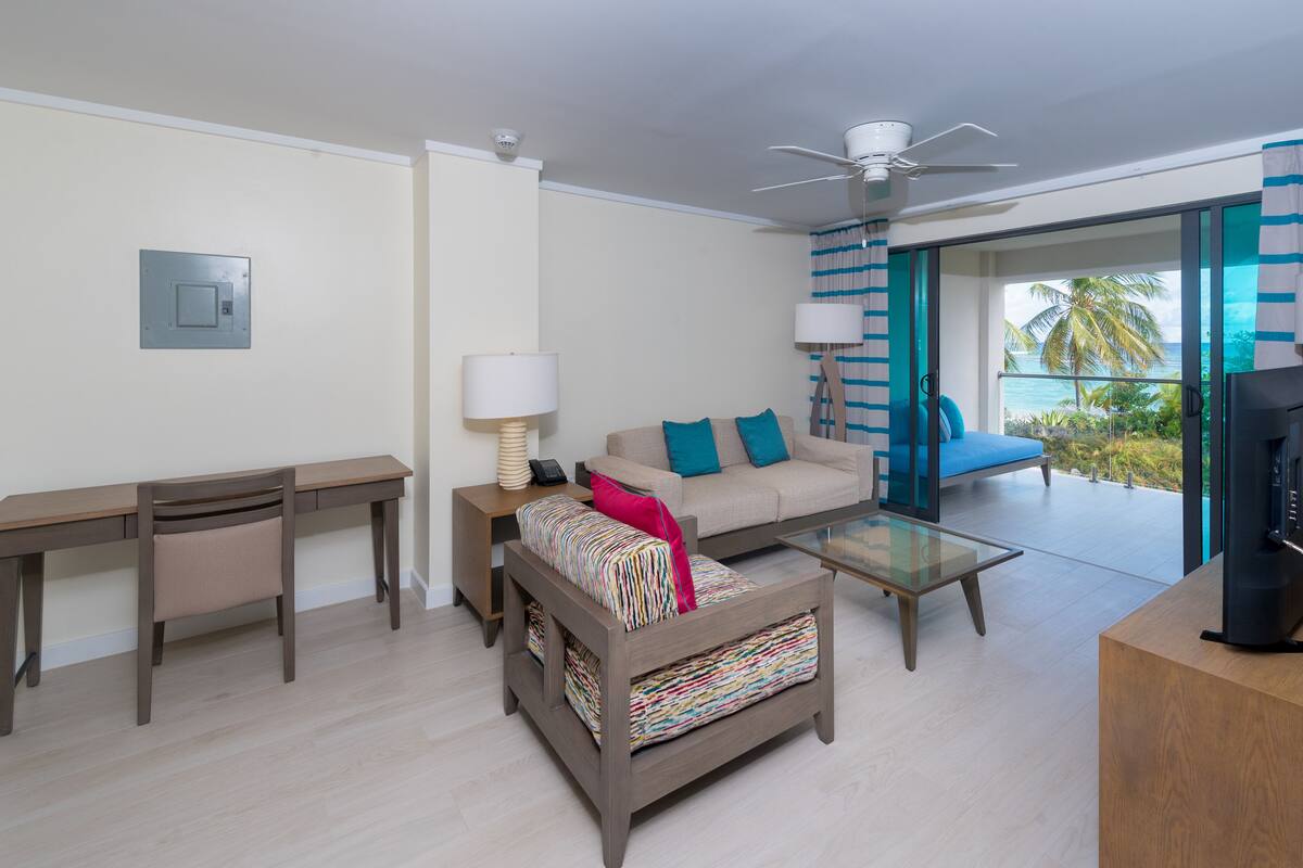 A modern living room with a sofa, coffee table, and a desk. There is a balcony with a view of the ocean.