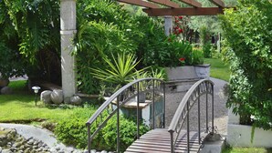 A garden with a metal bridge over a small stream, surrounded by various plants and a pergola structure.