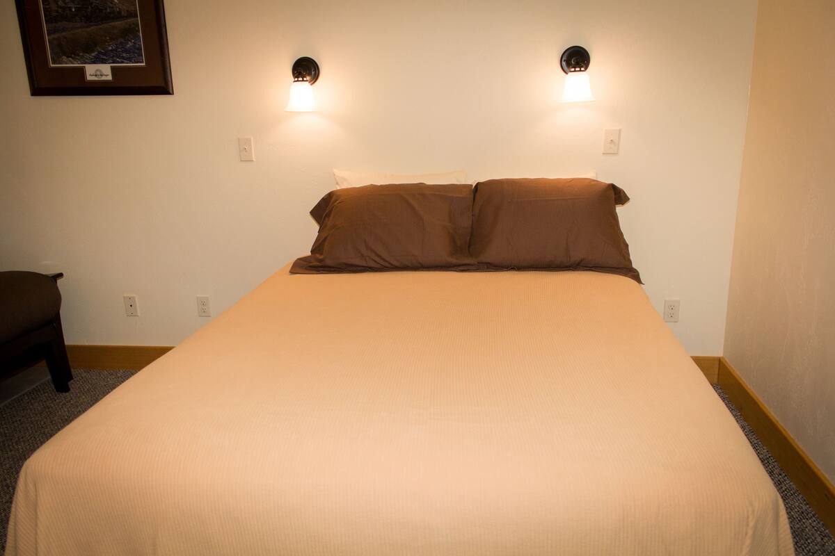 Un lit bien fait, recouvert d’une couvre-lit beige et agrémenté de deux oreillers, ainsi que deux appliques murales et un tableau encadré accroché au mur.