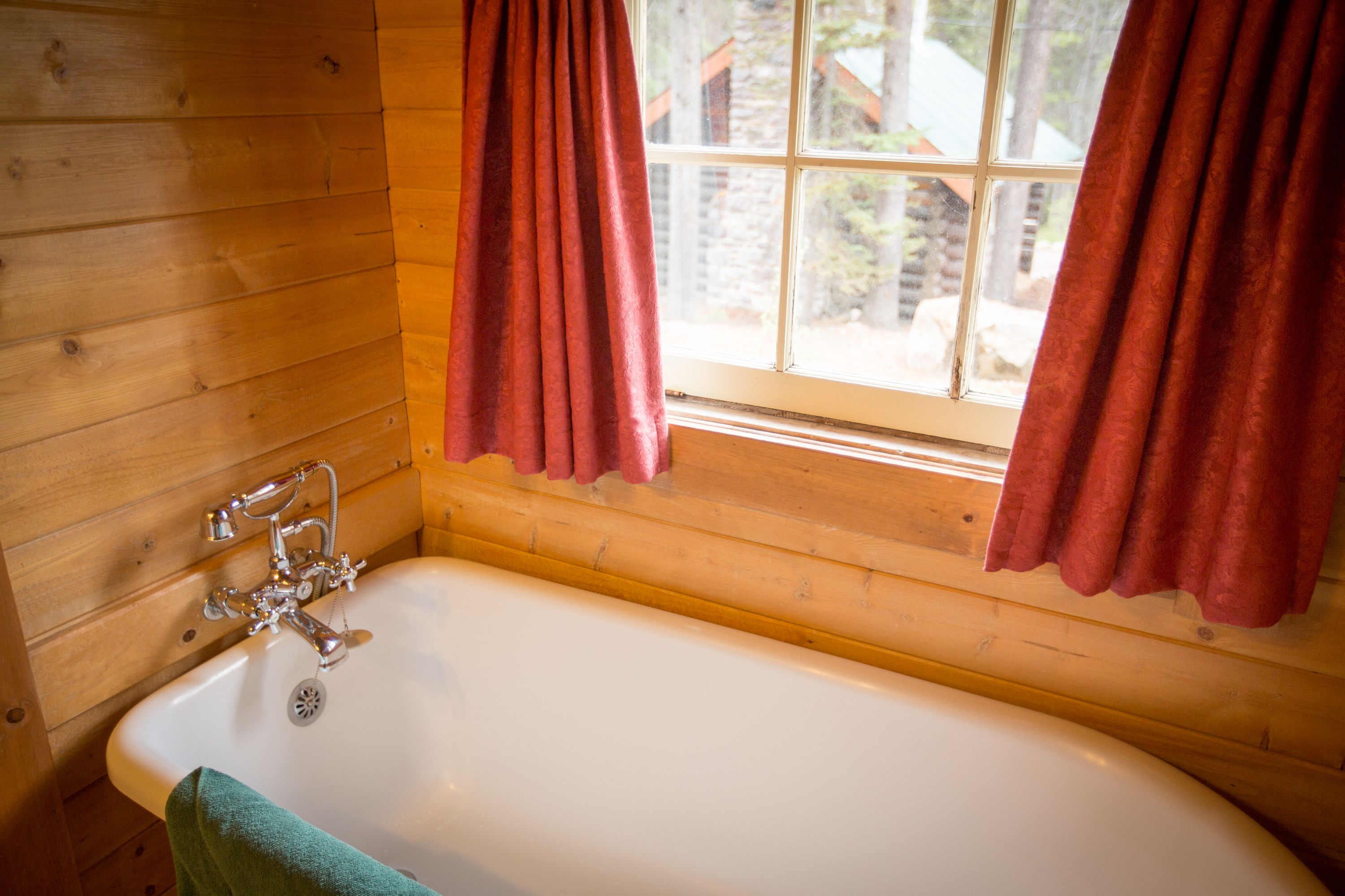 A wooden cabin bathroom with a white bathtub, a window with red curtains, and a green towel on the floor.