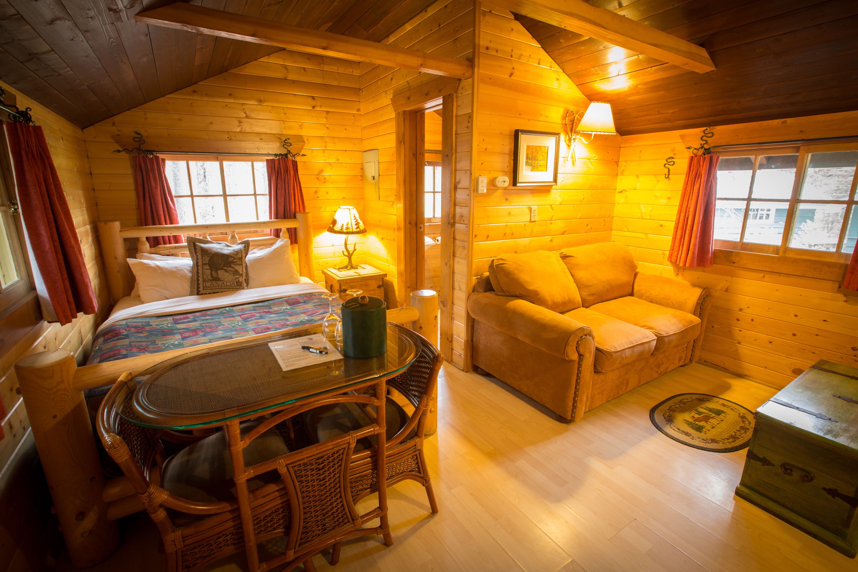 A cozy log cabin room with a bed, a sofa, a dining table with chairs, and a small table with a lamp.