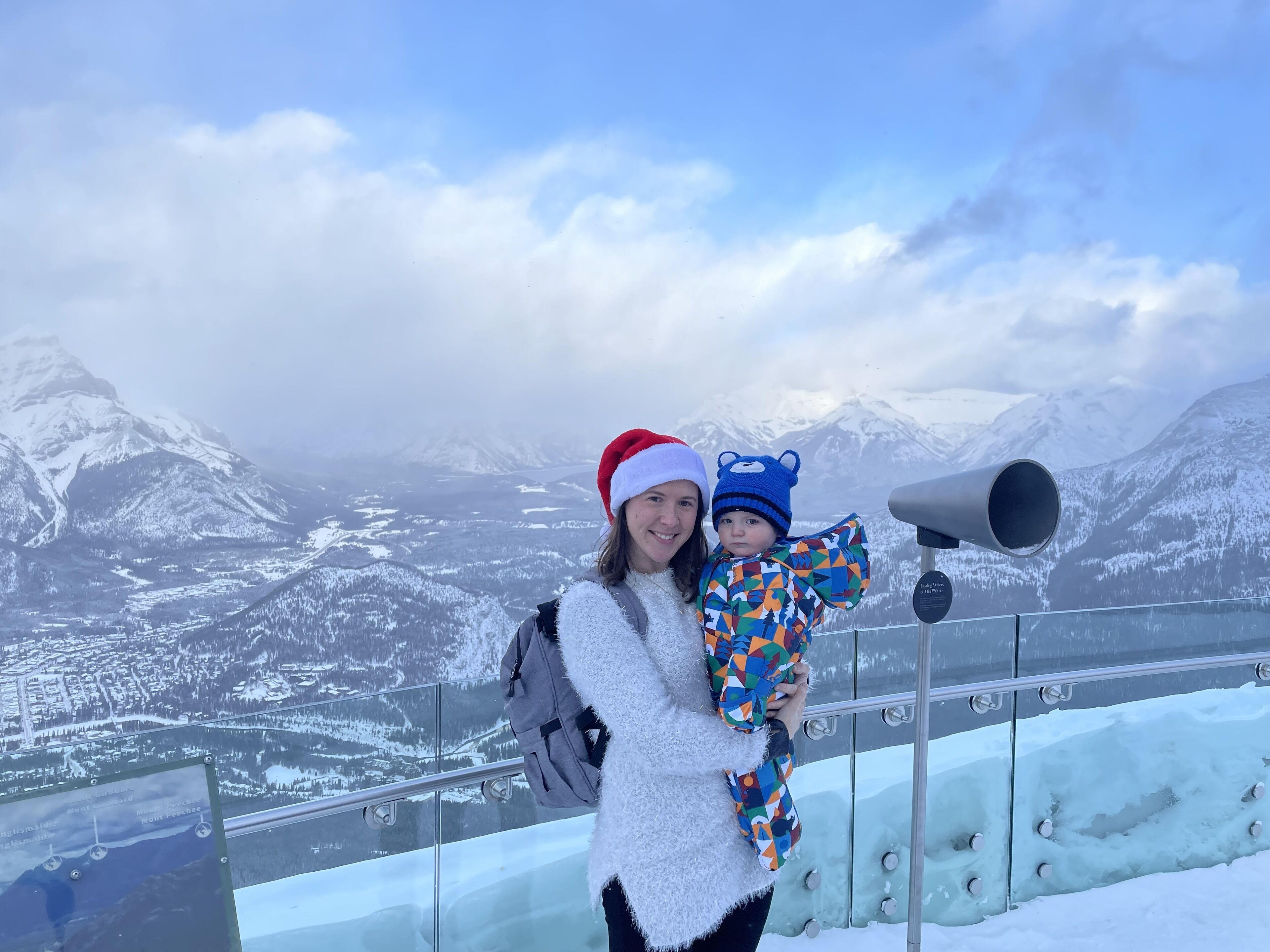 A person holding a child with a telescope, overlooking a snowy mountainous landscape.