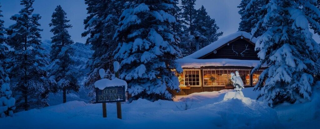 A lodge entrance sign in a snowy forest with a cabin in the background.