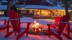 A cozy outdoor seating area with red Adirondack chairs around a fire pit, set against a wooden cabin with a snow-covered roof. - Storm Mountain Lodge Cabins & Dining (Castle Junction)