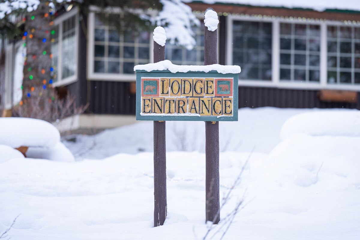 Letrero que dice 'Lodge Entrance' con techo nevado y postes de madera, frente a un edificio con ventanas y un árbol con luces de colores.
