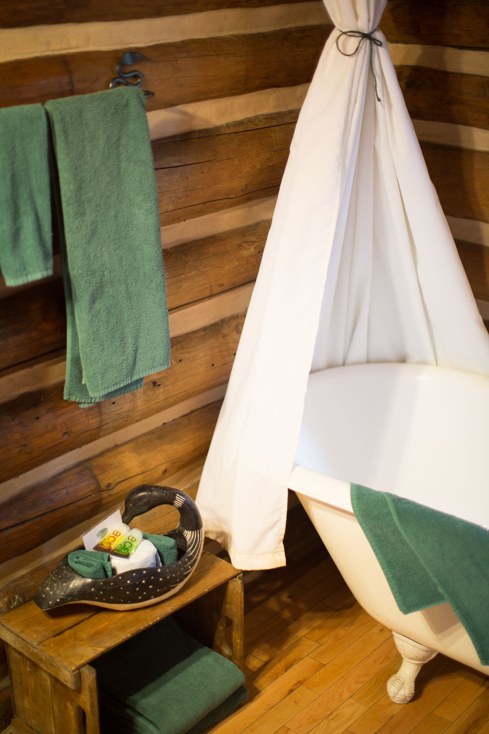 A rustic wooden cabin interior with a bathtub, a wooden stool with a green towel, and a green towel hanging on the wall.