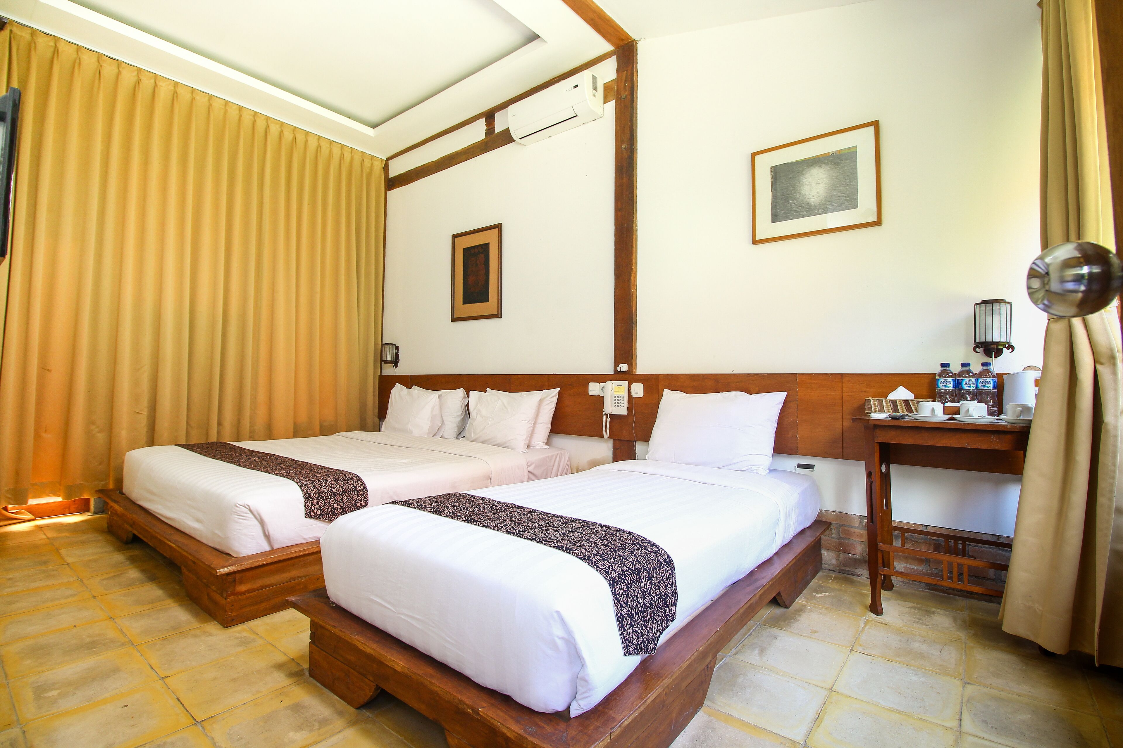 A hotel room with two beds, a wooden headboard, a bedside table with a lamp, a framed picture on the wall, and a telephone.