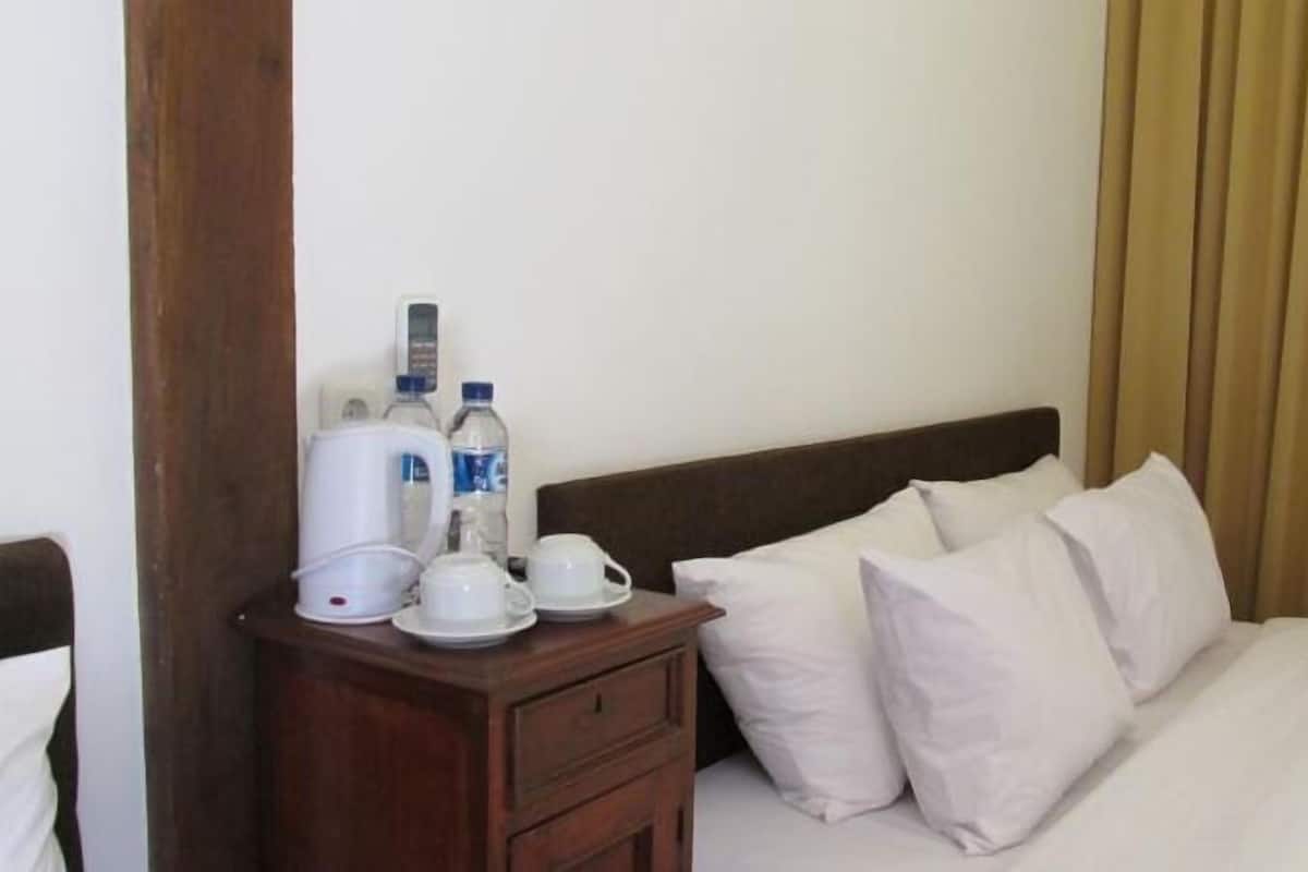 Habitación de hotel con cama, almohadas blancas, mesita de noche de madera y cafetera, tazas y botellas.