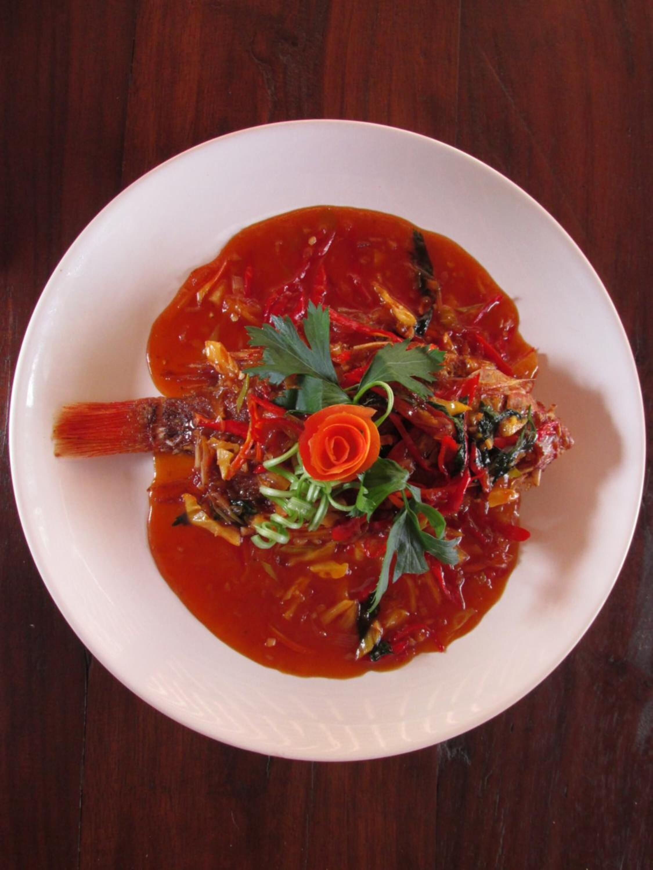 A white plate with a fish dish garnished with herbs and a decorative orange flower, served on a wooden table.