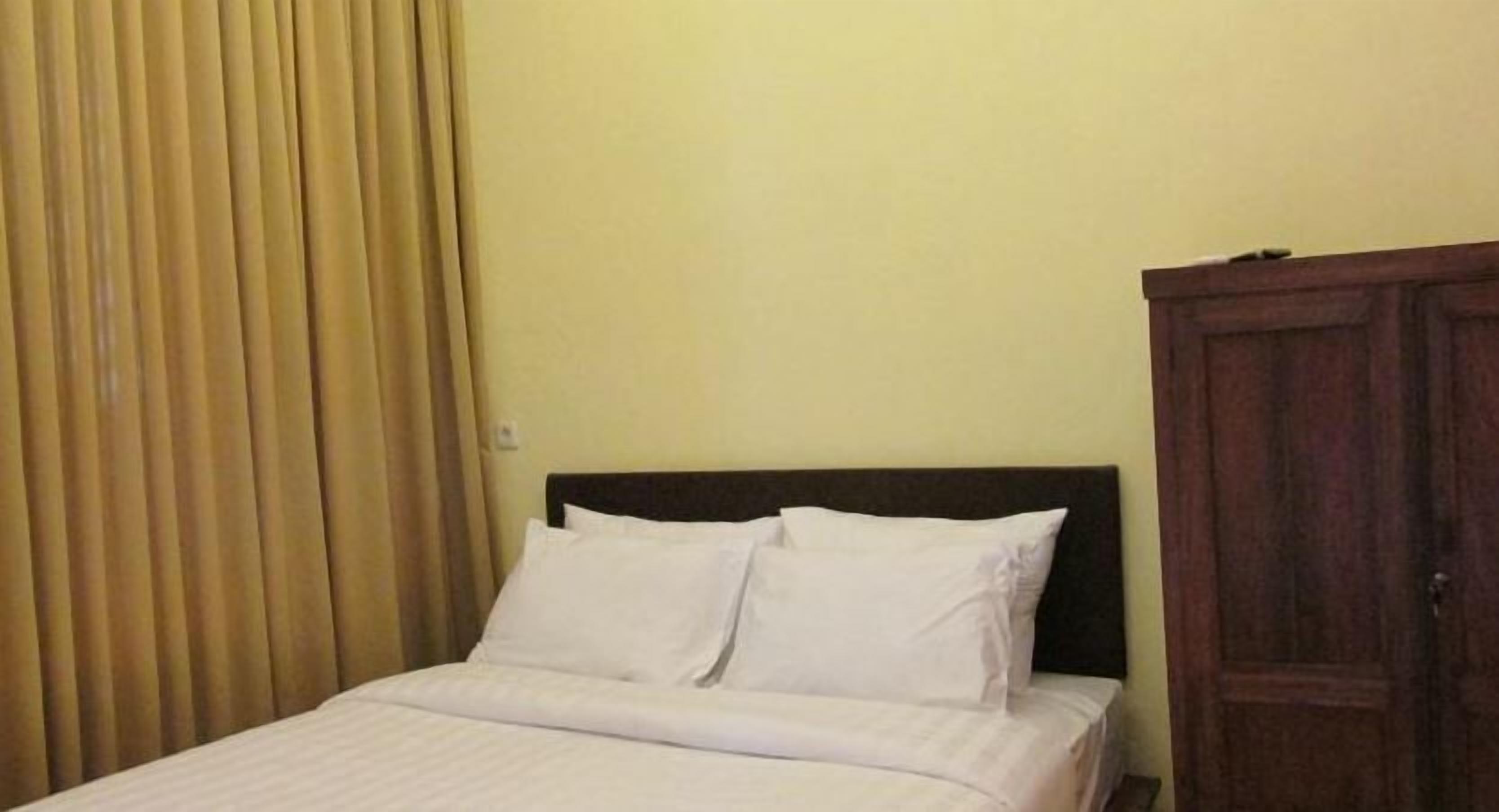 A neatly made bed with white linens and a dark headboard in a room with a wooden wardrobe and a beige curtain.