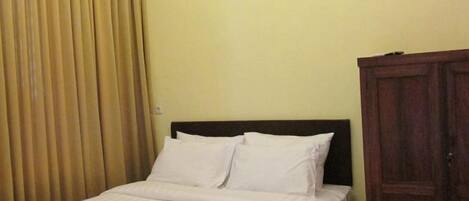 A neatly made bed with white linens and a dark headboard in a room with a wooden wardrobe and a beige curtain.
