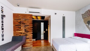 Quarto de hotel moderno com parede de destaque em madeira, cama com roupa de cama branca, área de estar e corredor que leva a outro quarto.