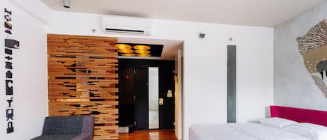 A modern hotel room with a wooden accent wall, a bed with white bedding, a seating area, and a hallway leading to another room.