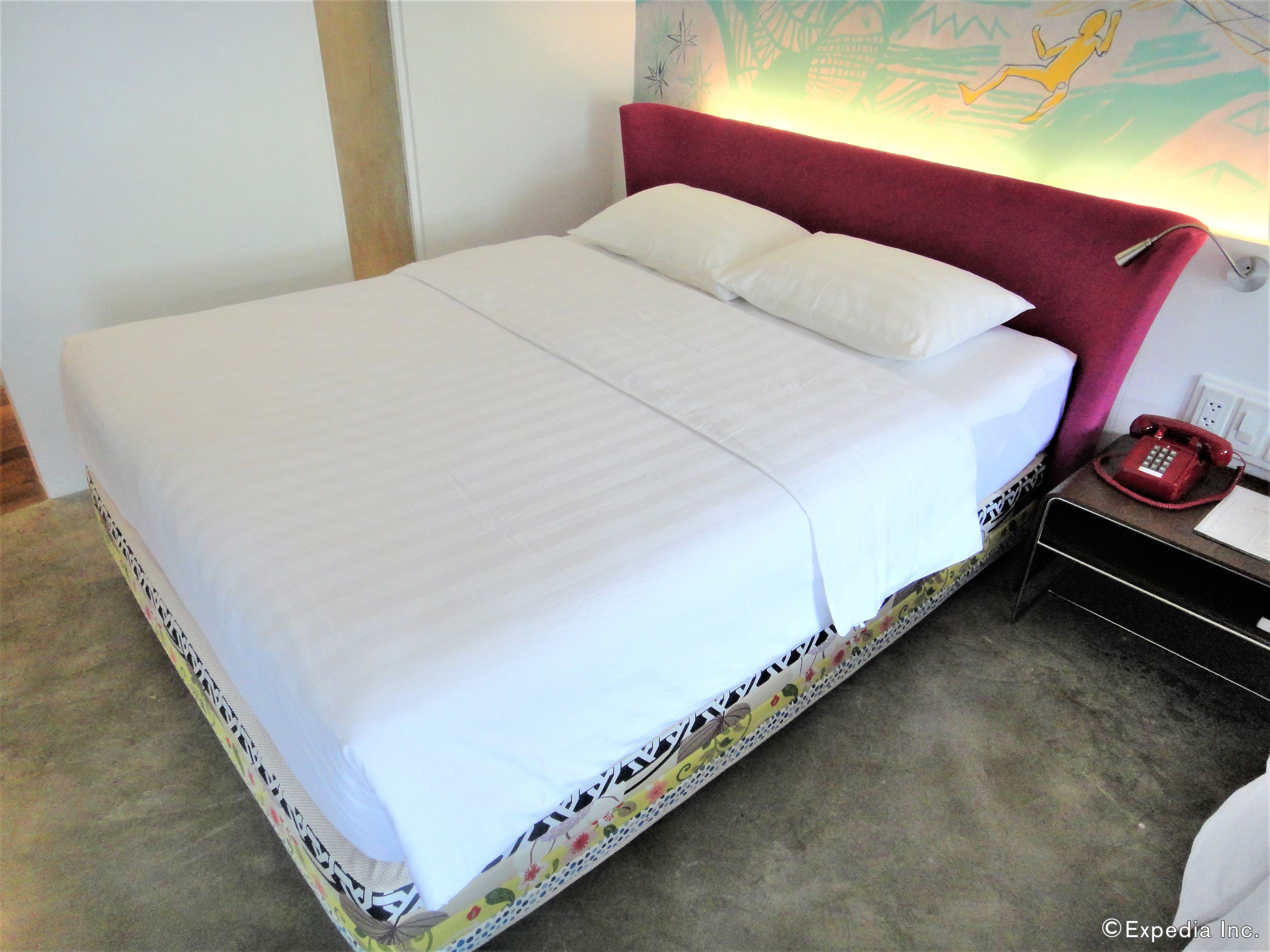 A neatly made bed with white linens, a red headboard, and a red telephone on a bedside table.