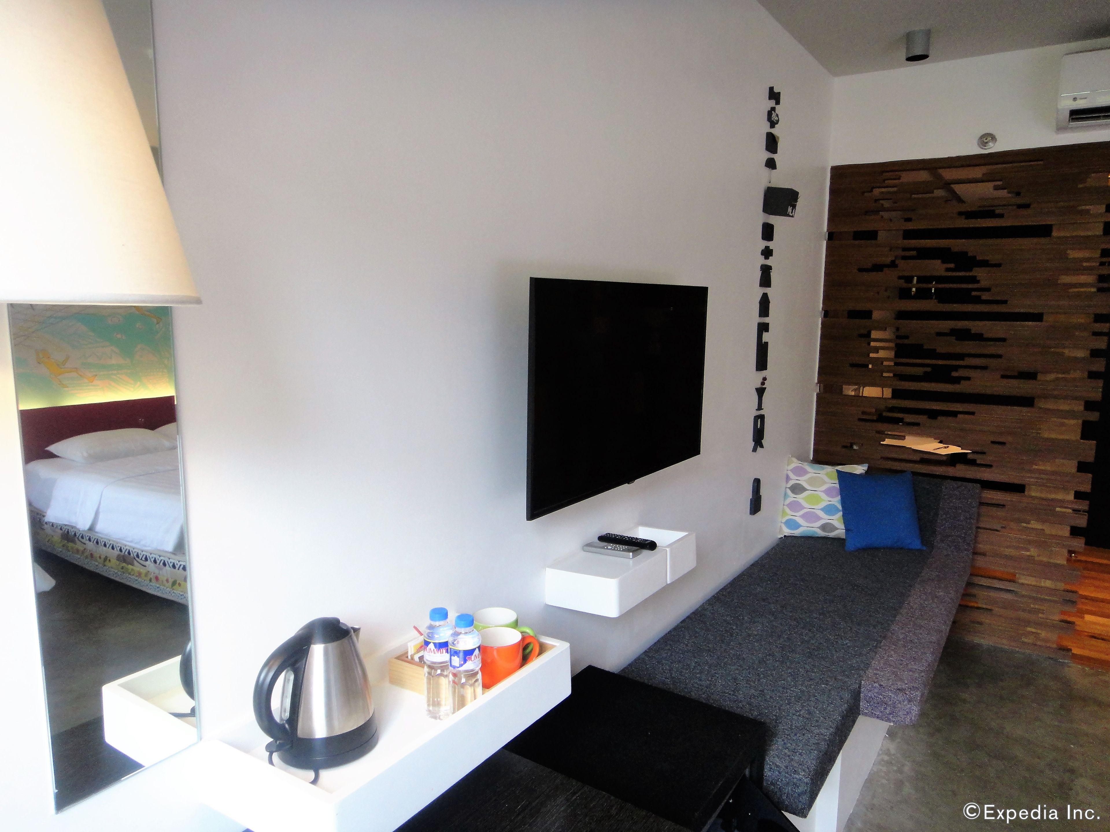 A modern hotel room with a flat-screen TV mounted on the wall, a bed with a headboard, a small table with a kettle, bottles, and cups, and a wooden accent wall.