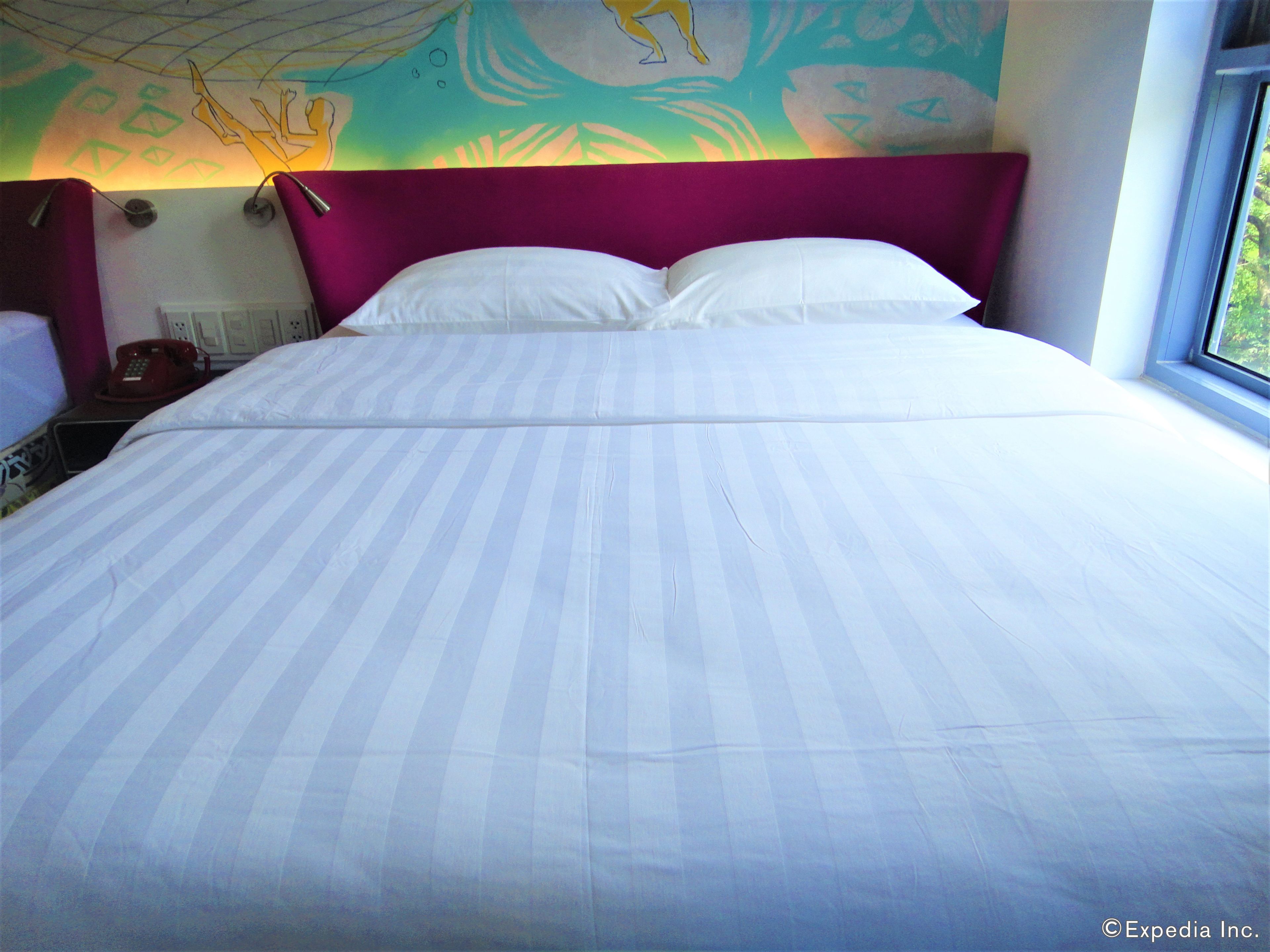 A bed with white and blue striped bedding, a purple headboard, and a colorful abstract mural on the wall behind it.