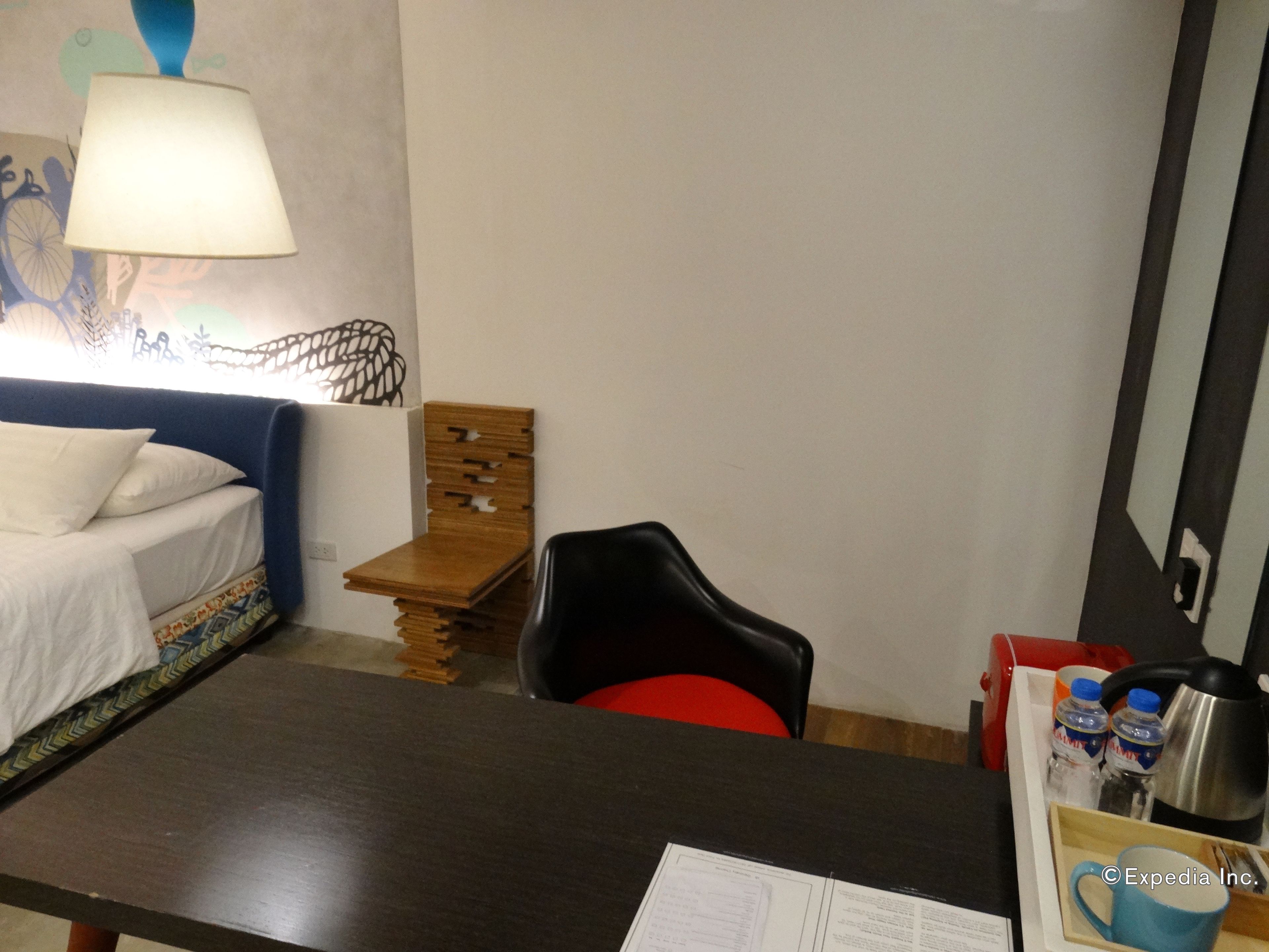 A hotel room with a bed, a desk, a chair, a lamp, and a wall with a decorative pattern.