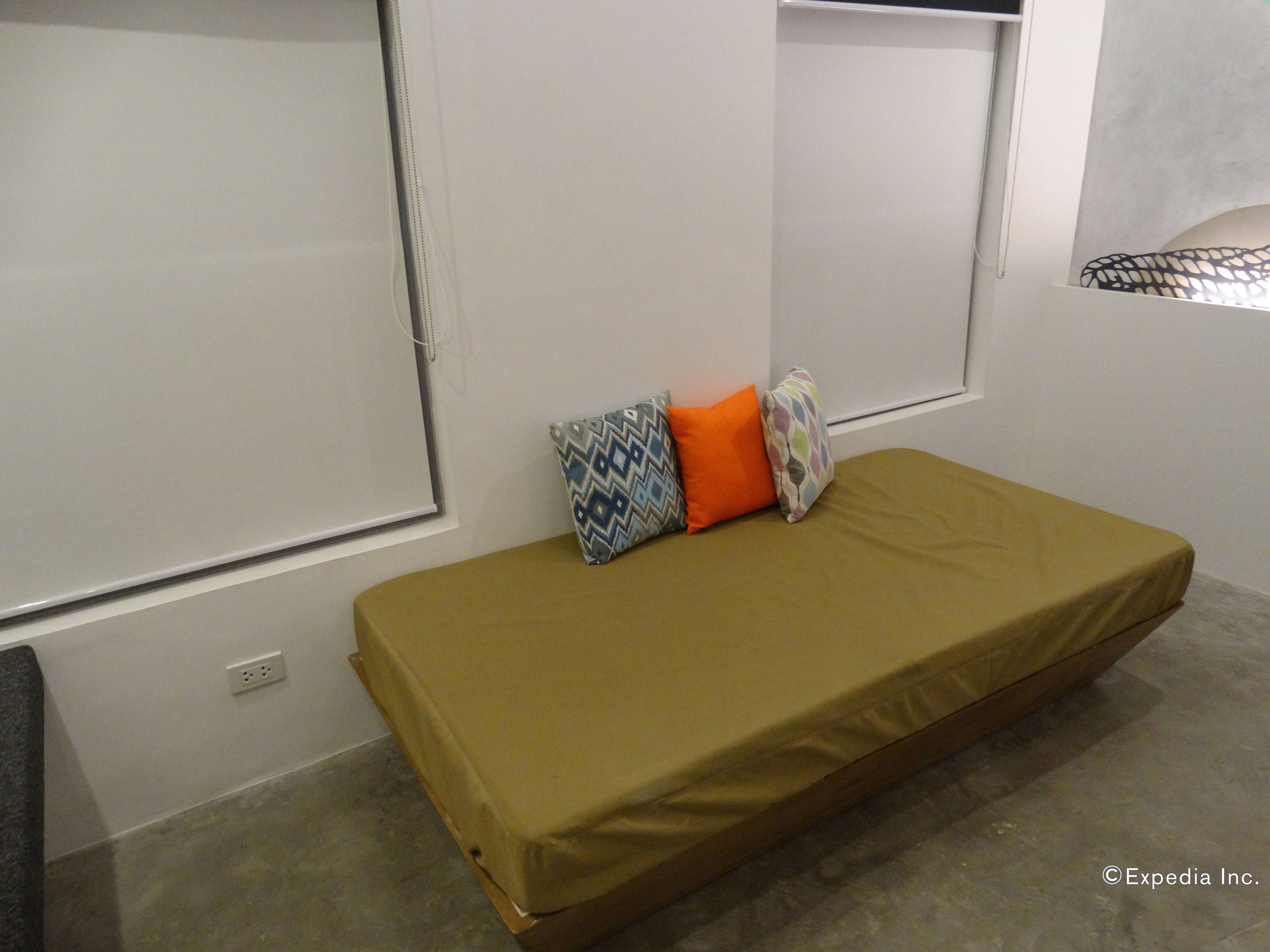 Una habitación pequeña con una cama individual, un colchón sencillo, una ventana con persianas y un enchufe en la pared.