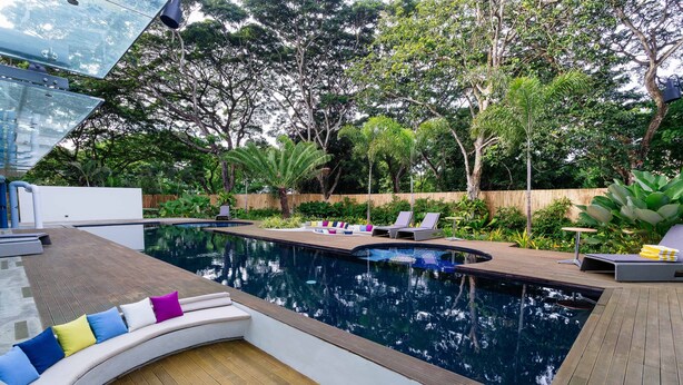 Uma área de piscina moderna ao ar livre, com deck de madeira, espreguiçadeiras e um espaço de estar curvo com almofadas.