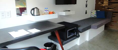 A modern kitchenette with a built-in oven, a microwave, and a coffee machine. There is a flat-screen TV mounted on the wall, a small shelf with a kettle and a water bottle, and a seating area with cushions.