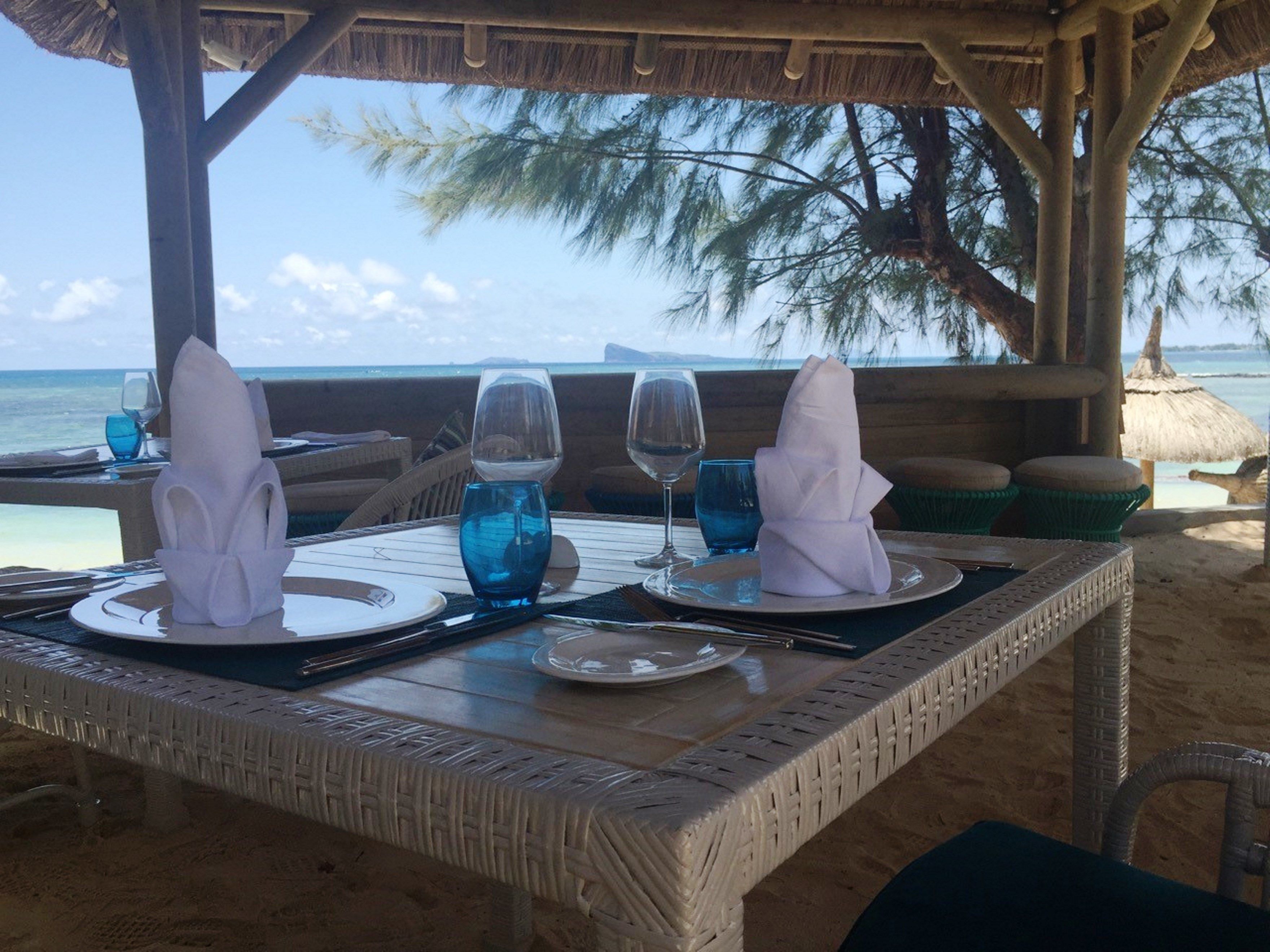 A table set for two with a view of the ocean, a thatched roof, and wicker furniture.
