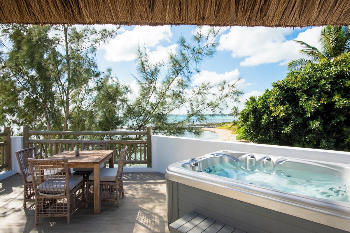Une terrasse en bois avec un jacuzzi, des fauteuils en osier et une table offrant une vue sur la plage.