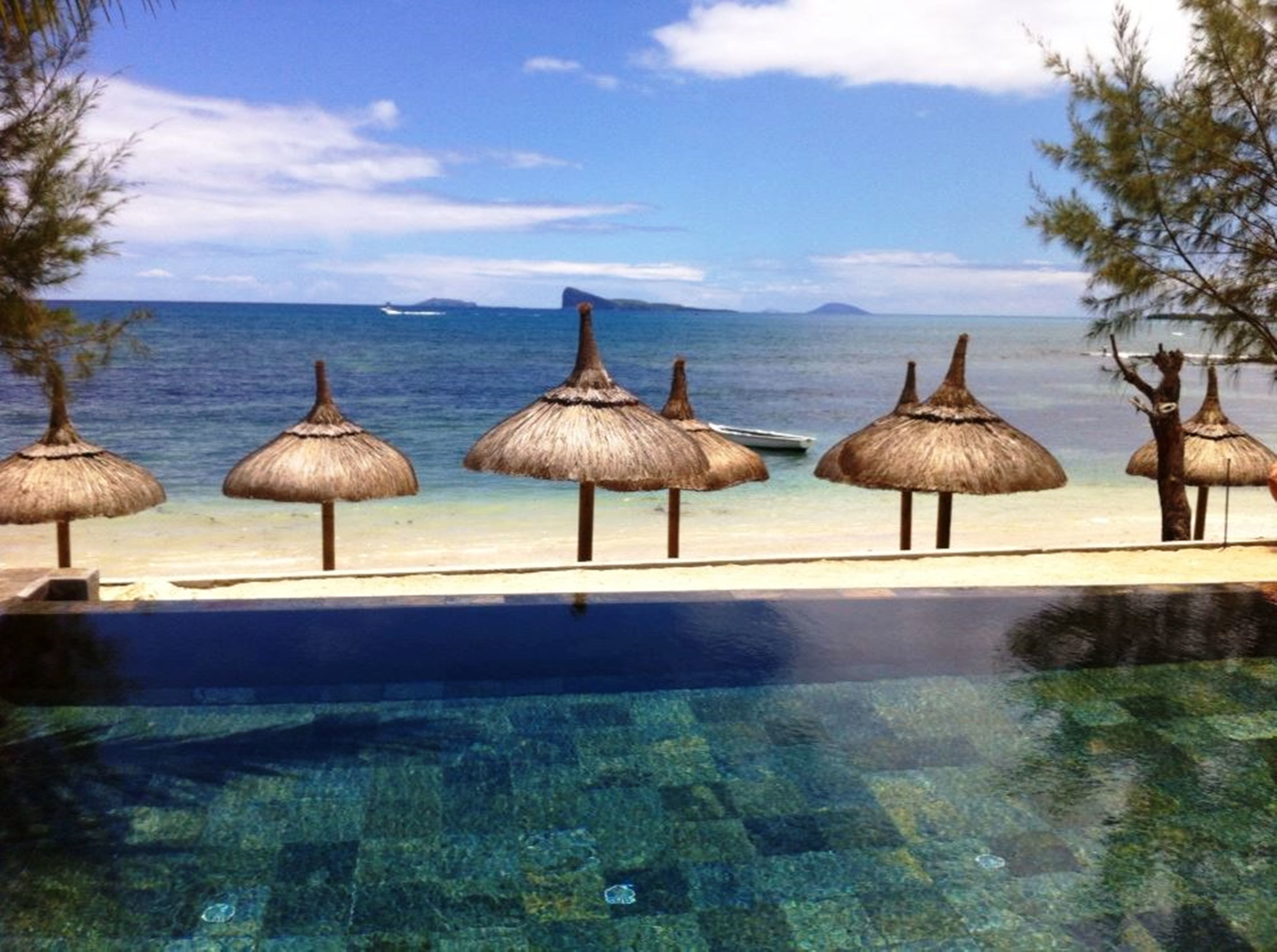 An infinity pool with clear water, surrounded by thatched umbrellas, a sandy beach, and a view of the ocean and distant island.